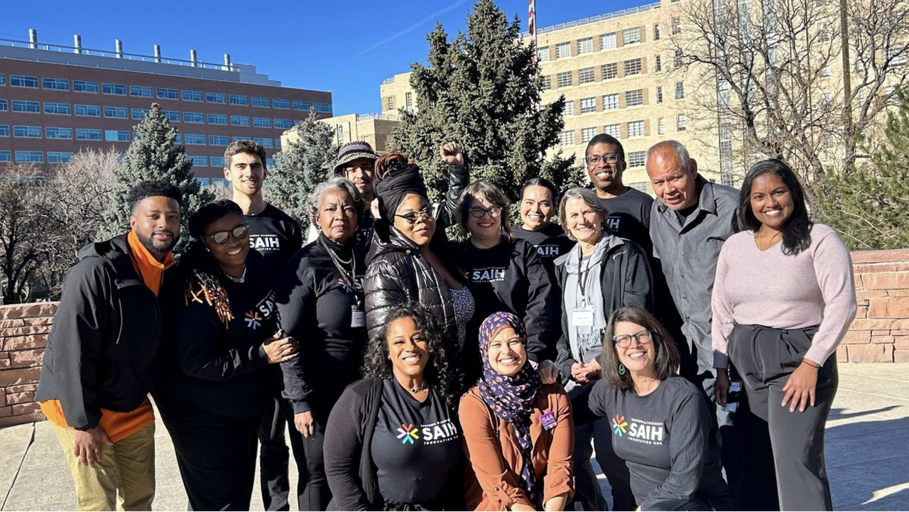 A group of Black, Brown, Indigenous, and white partners smiling in their SAIH t-shirts on the University of Colorado campus.