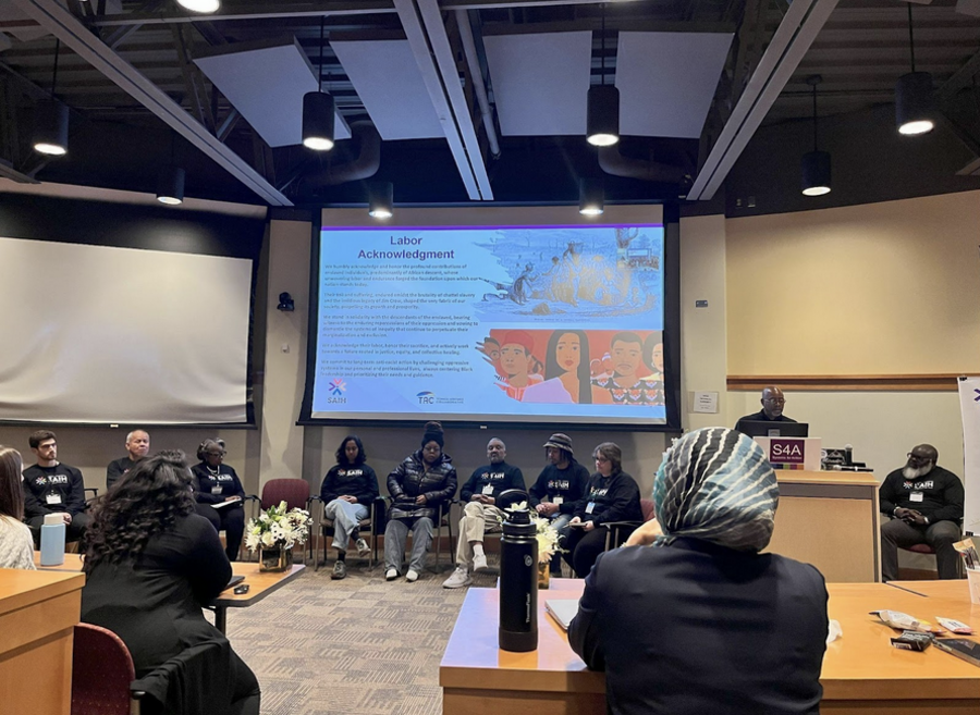A panel discussion in a lecture room, with seated SAIH grantees facing an audience while one of them presents. Behind the panelists is a projection of a written Labor Acknowledgement.